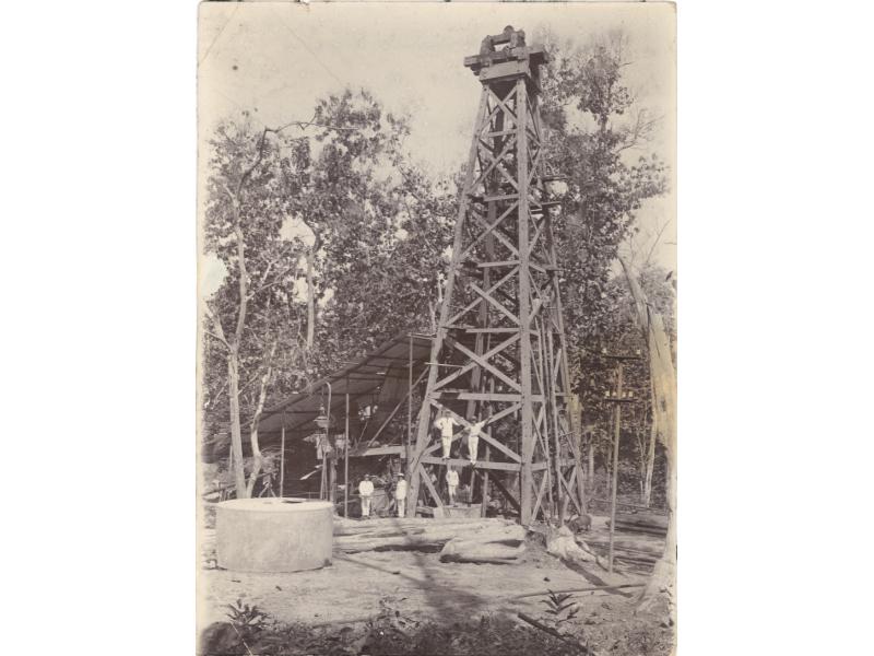 A photo of a group of International Drillers standing at the base and on the first support beam of an oil derrick. They are wearing white. There is a large cylinder and some logs in front of them and trees in the background.