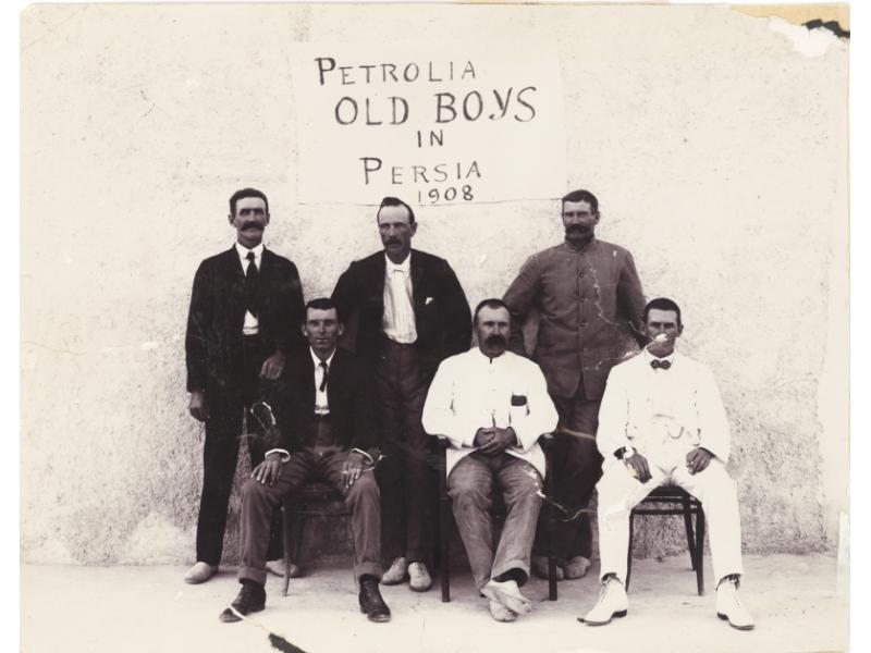 Six hommes en costume. Trois sont debout et trois sont assis. Une affiche sur le mur derrière eux indique « Les vieux garçons de Petrolia en Perse, 1908 ».