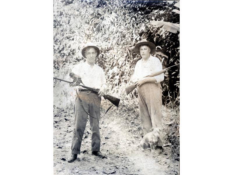 Two men in white shirts, hats, and dark pants.They are holding rifles. There is a small dog at the foot of the man on the right. They are standing in front of a wall of vegetation.