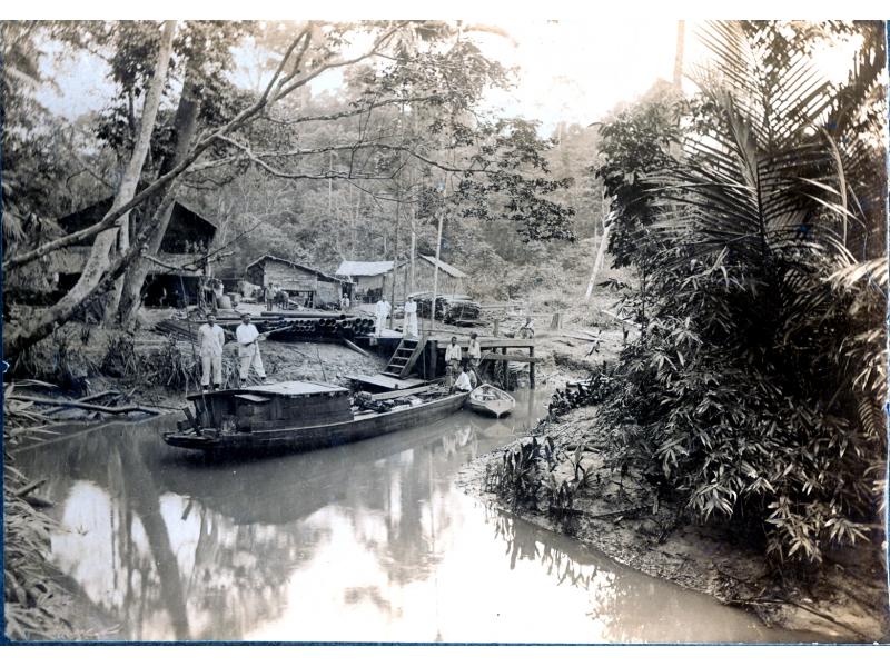 Six men on a boat in a narrow river. There are two men on a dock with a pile of casing beside them on the shore. A group of men stands behind the basing in front of three buildings. There are trees in the background.