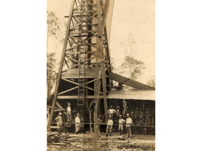 Un groupe d'hommes debout devant une installation de forage pétrolier en bois. Il y a une échelle à l’avant de l’installation. À côté de l’installation, la machinerie est couverte par un toit en tôle.