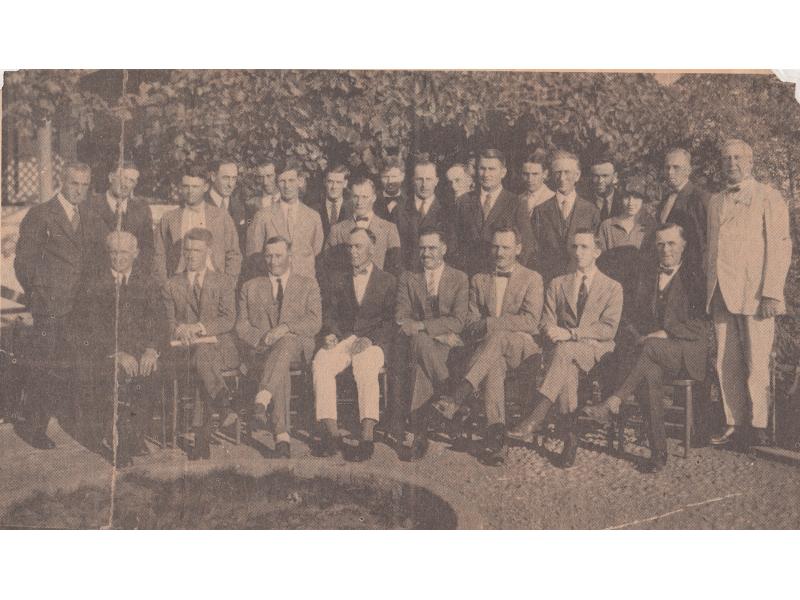 A group of men in two rows. One row is standing and the other is sitting in chairs. They are wearing suits and are in front of large bushes.