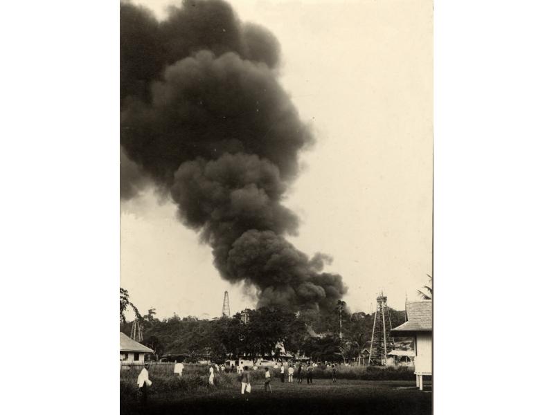 A photo of a group of people watching a cloud of black smoke near a group of oil derricks.