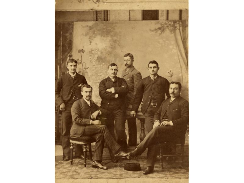 A photo of six men in front of a painted backdrop. They are wearing dark suits and two are sitting in chairs. There is a patterned rug underneath their feet