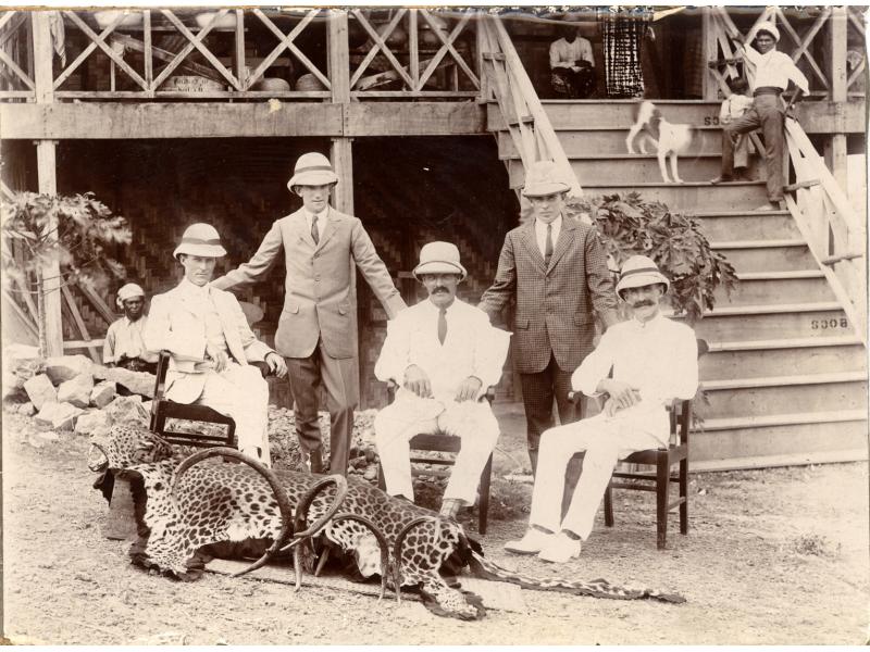 A photo of five men wearing pith helmets, two standing and three sitting in chairs, behind a large cat skin and two sets of animal horns. There are two people and a dog on a set of stairs leading up to a building behind the men.