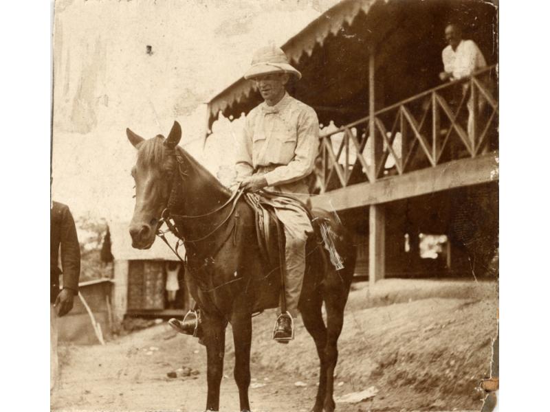 Une photo d’un foreur international vêtu de blanc, coiffé d’un casque colonial et assis sur un cheval. Il est devant un bâtiment monté sur pilotis.