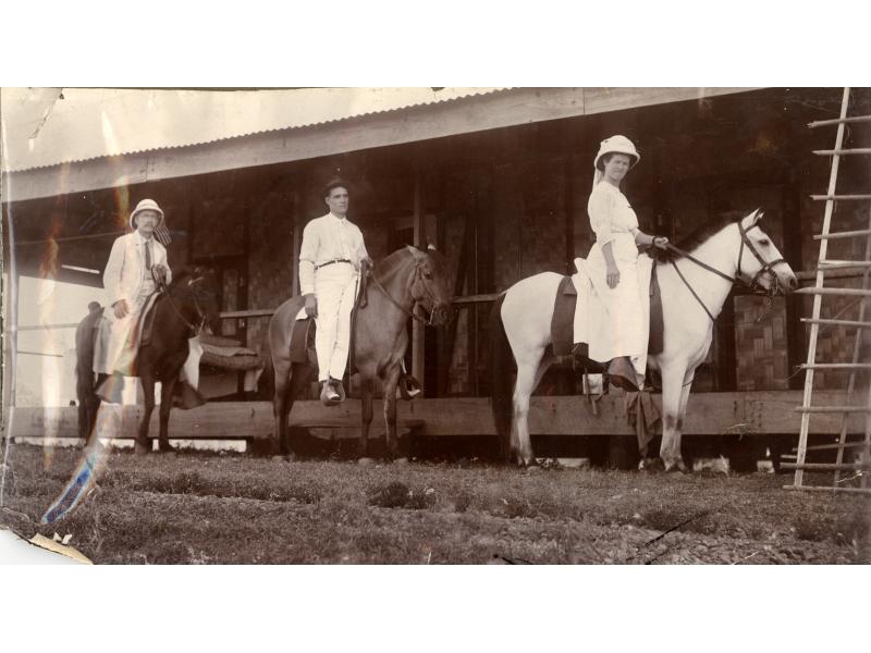 Two men and a woman on horseback in front of a building with a raised deck. The men are riding dark horses and wearing white shirts and pants. The woman is wearing a white dress and hat. She is riding a white horse.