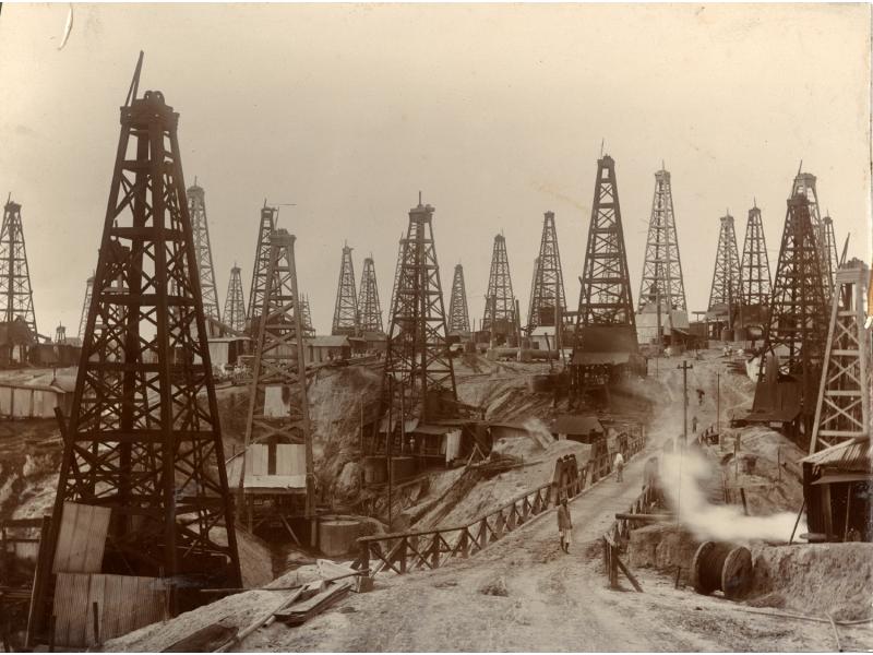 Close to thirty oil derricks with four poles each. There is a bridge leading across a low point in the oil field and several men are walking along the road. There are buildings behind the front row of derricks and a large groups of people are gathered near a pile of casing.