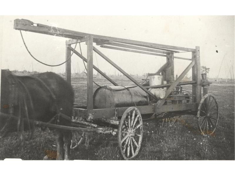 A horse harnessed to a crude oil dipping machine. It is a cylindrical tank on a cart with four wheels. There is a wooden beam folded forward on top.