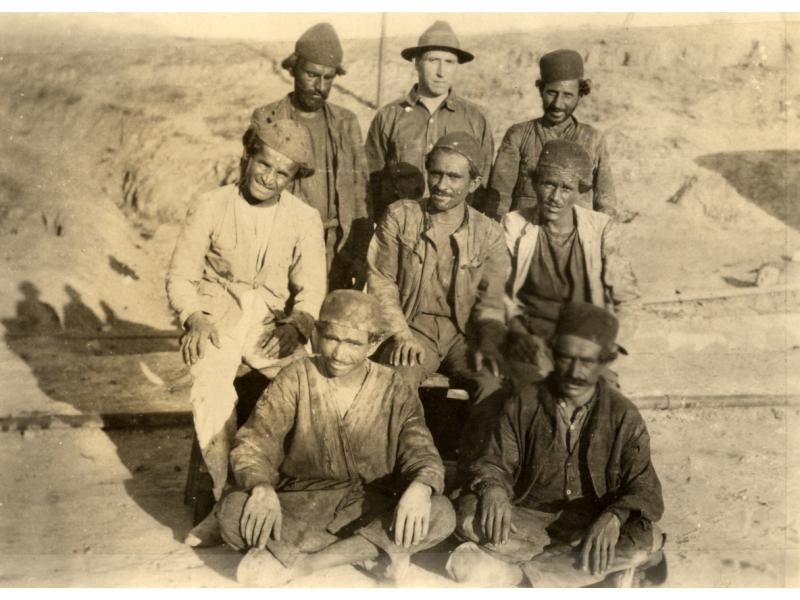 Charles Warren stands with seven local men arranged in three rows, all wearing work clothes. Warren is in the centre in the back row. The middle row is sitting on chairs and the front row is sitting cross-legged on the ground.