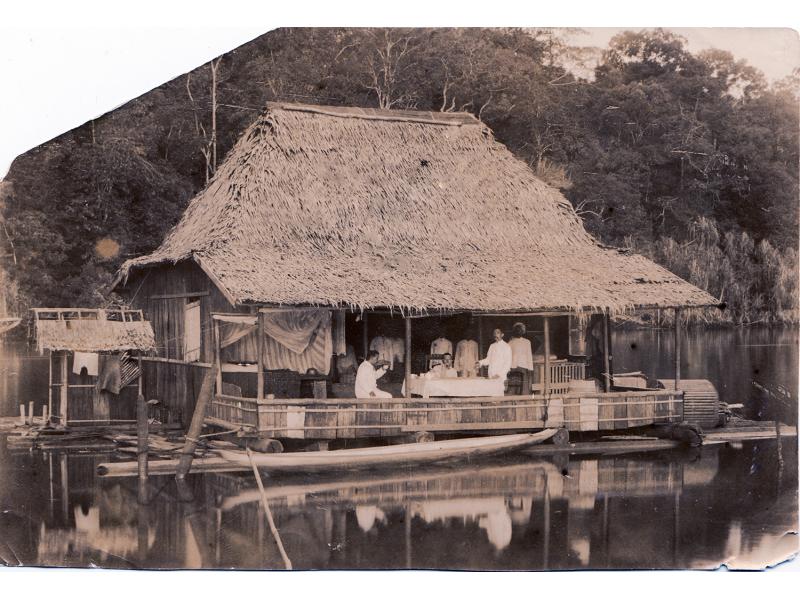A group of men sit on the verandah of a straw-thatched bungalow on the bank of a river. 