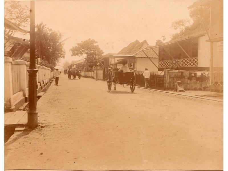 A view of a street in Palembang with a few carts and people walking under umbrellas against the sun.