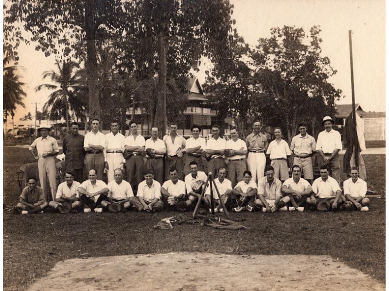 A baseball team. Half the men are standing and half are sitting. Most are wearing white t-shirts. They are in front of a group of trees and a building. There are three baseball bats set up like a tripod in front of them with a ball resting on top. Some other equipment is also in front of them.