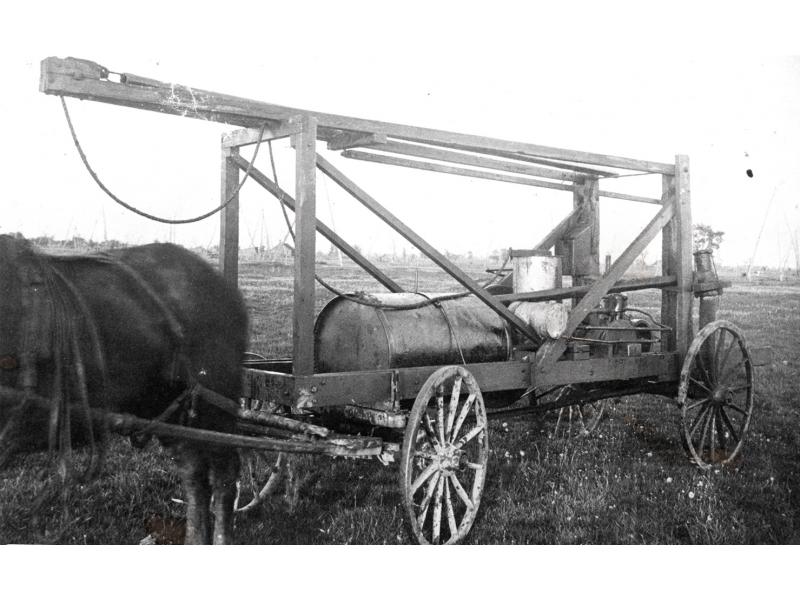 A horse pulling a cart with four wheels. There is a wooden beam sticking out over an open wooden frame that has a metal barrel resting in it.