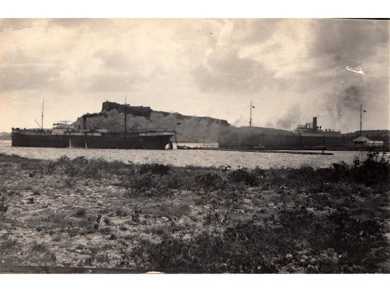 A photo of a tanker tied to a dock along a rocky shoreline. There are four pipes running along the left side of the image and there are boats out in the water in the background.