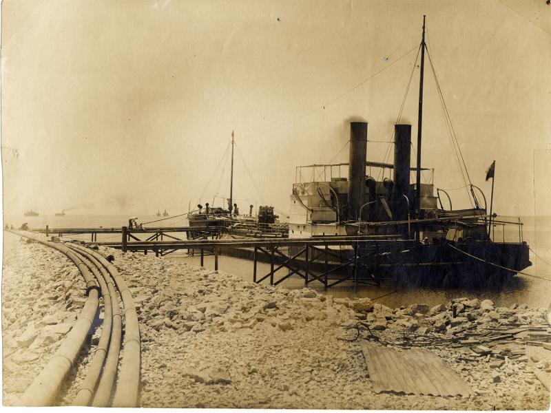 Photo d'un pétrolier amarré à un quai le long d'un rivage rocheux. Quatre tuyaux longent le côté gauche de l'image et des bateaux se trouvent dans l'eau, à l'arrière-plan.