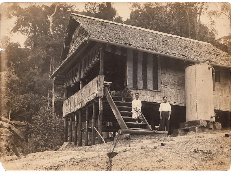 A thatched bungalow on stilts. A woman and a dog sit on the steps leading up to the porch and a man stands beside the stairs.