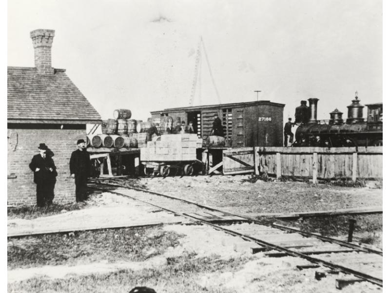 Two men stand outside a brick building. Oil barrels are being loaded onto a train. There is an three-pole derrick in the background.