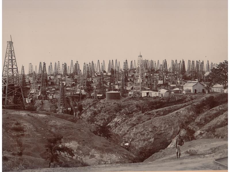 Over a hundred derricks standing in a field. There are buildings and storage tanks in between them. There is a man standing on a hill that looks across a small valley in front of the field.