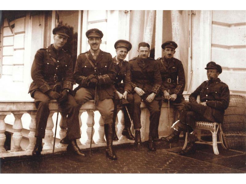 A photo of six men dressed in army uniforms and holding canes. They are sitting on a white railing in front of a building with white siding. The man on the right is sitting on a wicker chair.