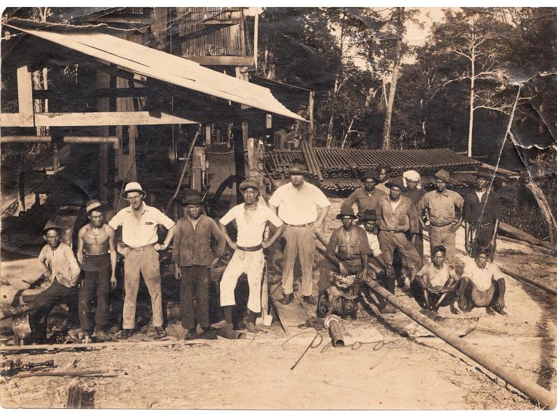 Group of fourteen local workers and two international drillers, all dirty and covered in oil stains, standing in front of a rig in Borneo. 