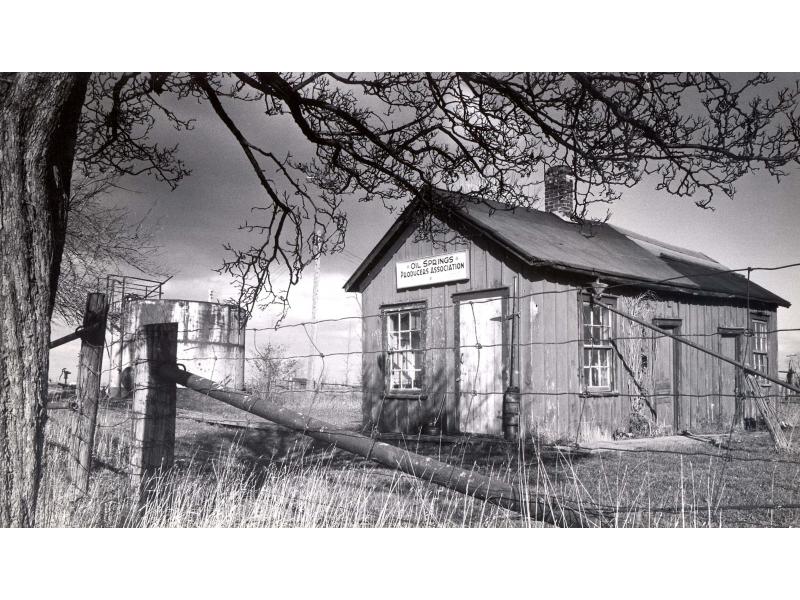 A small building with three windows and a white door behind a wire fence. There is a storage tank in the background.