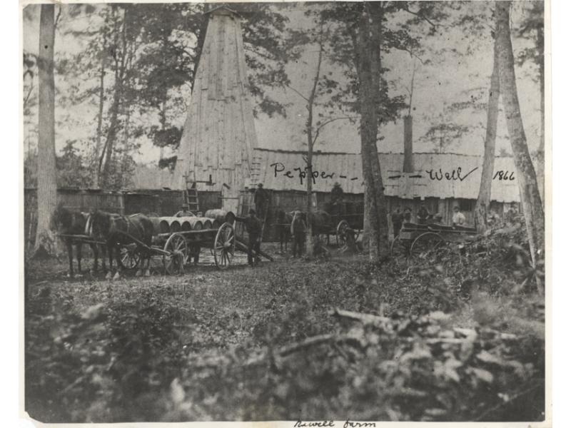 Deux équipes de chevaux devant des charrettes chargées de barils de bois, pendant que des hommes ajoutent d’autres barils. Il y a un grand bâtiment derrière eux, et « Puits Pepper 1866 » est écrit sur la photo.