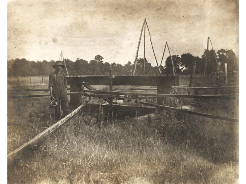 Un homme est debout près d’une roue de champs avec des rayons qui s’éloignent dans toutes les directions, et un derrick à trois mâts en arrière-plan. 