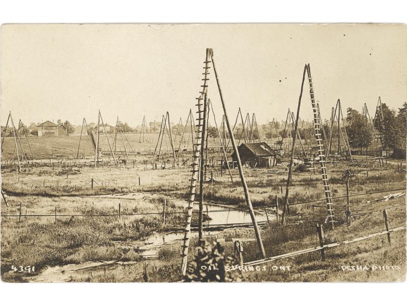 Three-pole derricks in a field with a creek running through it. There is a small building in the centre and buildings in the background.