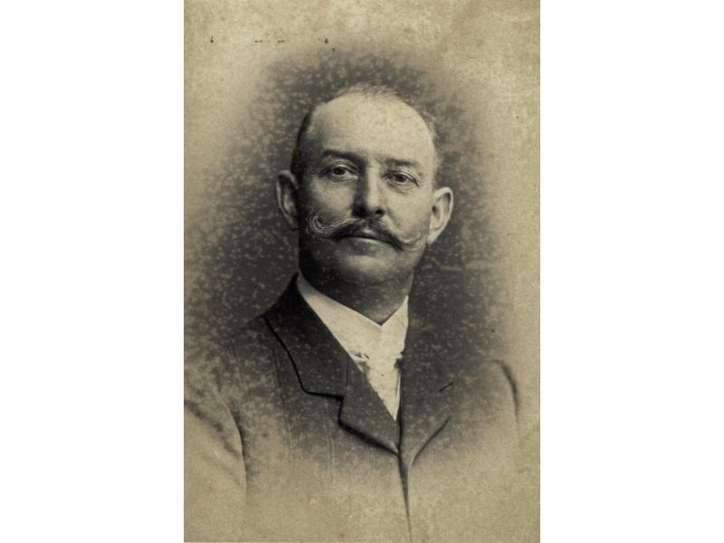A portrait photo of Jacob Perkins. He has a curled mustache and is wearing a dark jacket over a white-collared shirt.