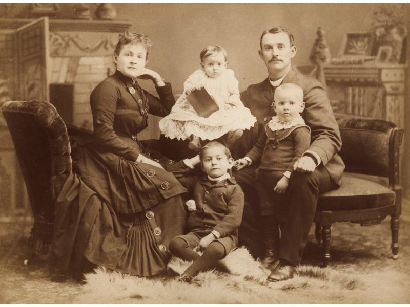 A woman and man sitting with three children between them. They are wearing dark clothing except for the youngest child in the middle who is wearing white. There is a mantle behind them and a fur rug at their feet.