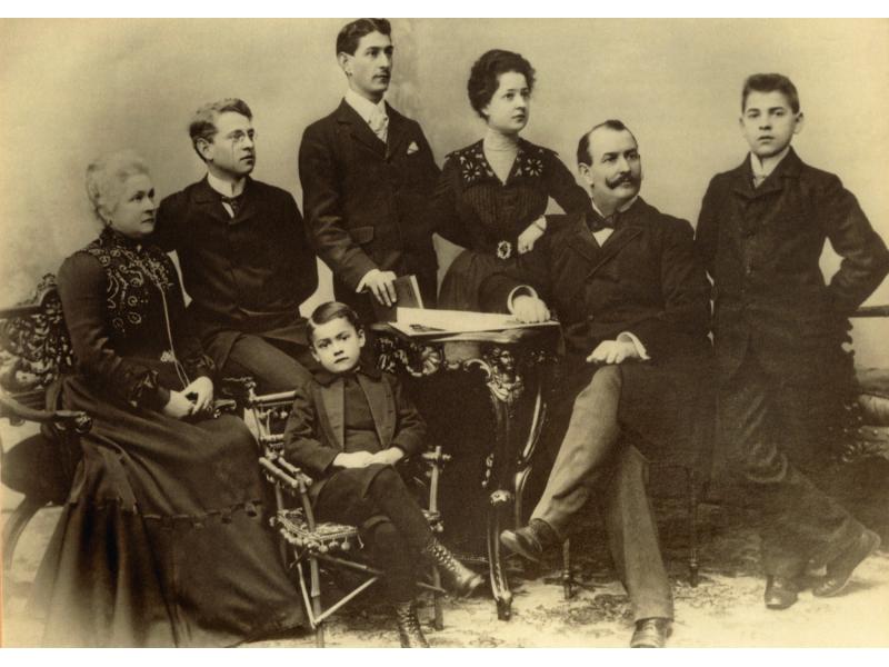 A posed photo of eight people, looking off to the right. They are wearing dark clothing. The two women (far left and third from right) have embroidery on the tops of their dresses.