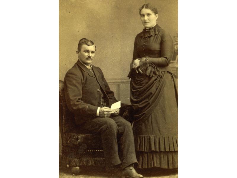 A photo of Charles and Jessie Nicklos. Charles is sitting in a chair and holding a piece of paper while Jessie is standing with her arms folded.