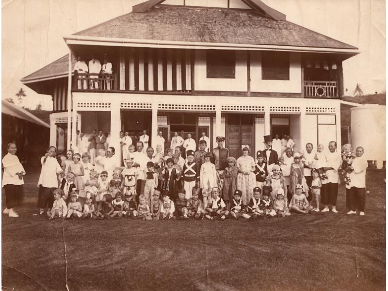  A group of children, some dressed as soldiers and angels, in front of what is probably a school. A group of adults stands behind on a deck.