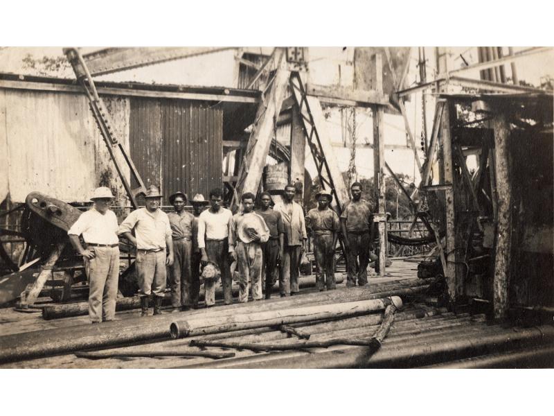 Two International Drillers wearing white and eight labourers in work clothes standing in front of a rig. There is a pile of casing in front of them.