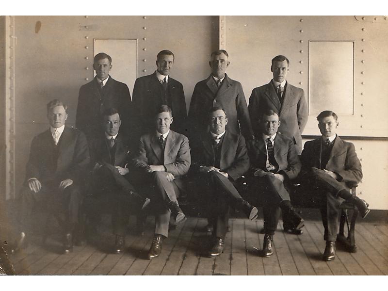 A photo of four International Drillers standing and six sitting in chairs on a boat deck. They are wearing dark suits and ties. They are standing in front of a wall that has five columns of rivets in it.