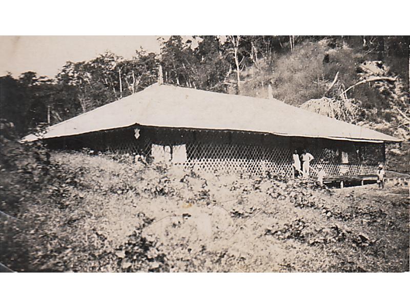 A bungalow set into a hill with a light roof and large latticework surrounding it. Three people stand on the stairs and another stands a few feet away.