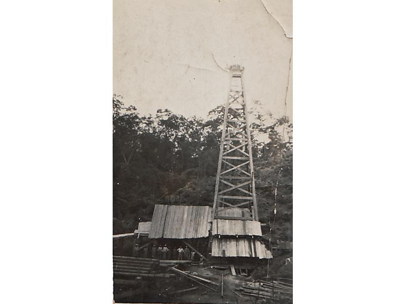 A photo of a covered oil rig. There is a group of men in front of it. There is casing in the foreground and trees in the background.