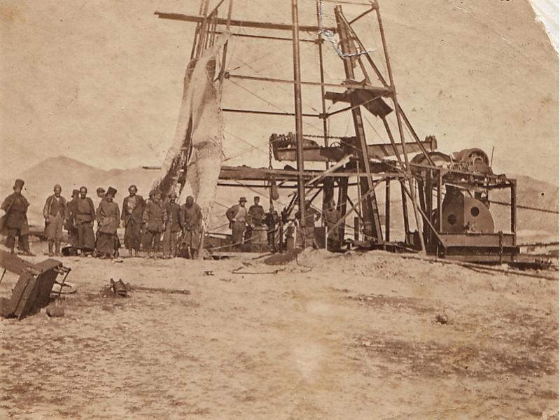A photo of a group of men wearing work clothes stand at the base of a metal oil rig. There is a piece of cloth covering the left side of the rig. The ground is bare and there are hills in the background.