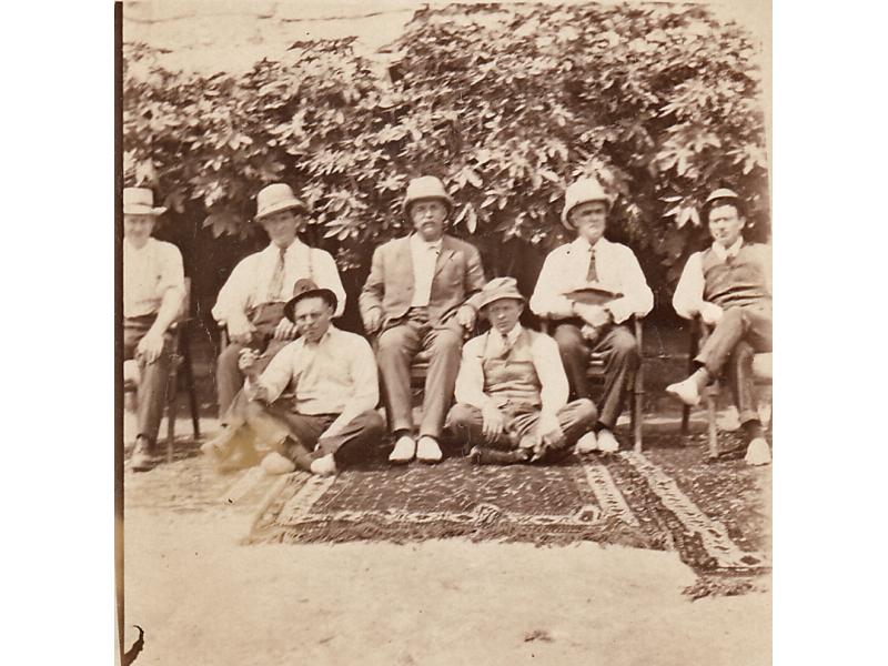 A photo of three patterned rugs on the ground.Five International Drillers are sitting in chairs and two are sitting on the ground. There are bushes behind them.
