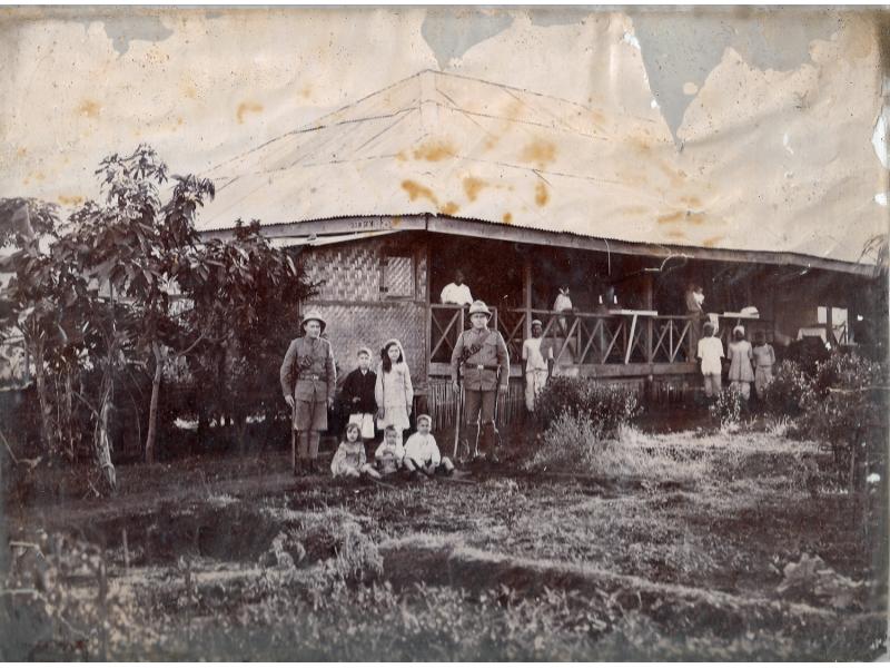 A photo of a thatched bungalow. There is a group of people outside the house, and two of the men are in uniform. There are trees on either side of the photo.