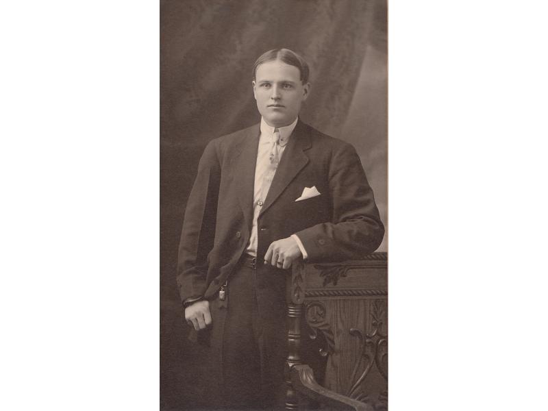 A portrait of Arthur Brown. He is leaning against the back of a carved chair. He is wearing a dark suit with a white shirt and tie. His hair is parted in the middle.