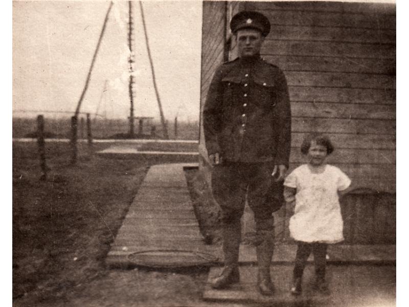 Arthur Brown standing next to Dorothy. He is wearing a military uniform and she is in a white dress. They are standing in front of a wooden building with wooden planks around it. There are derricks in the background.