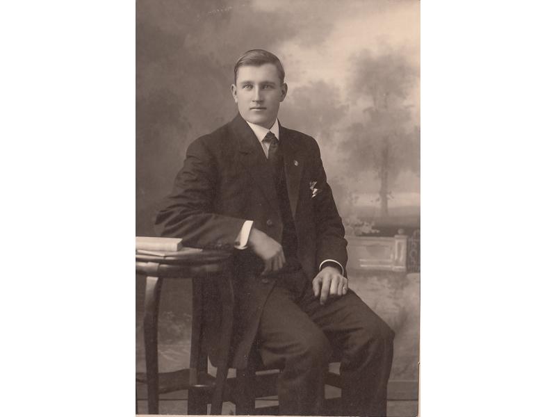  A photo of a man wearing a dark suit and tie, sitting in a chair and resting his elbow on a small table. There is a painted backdrop of trees behind him.