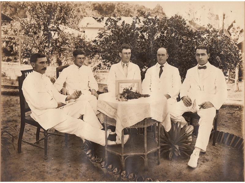 Five men sit around a wicker table covered with a white tablecloth. There is a bouquet of flowers in front of a frame on the table. The men are wearing white suits.