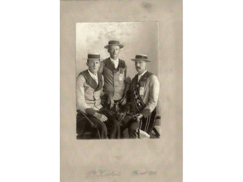 Three men wearing jackets and boat hats. The man in the middle is standing and the other two are sitting on chairs. The man on the right is wearing a sash.