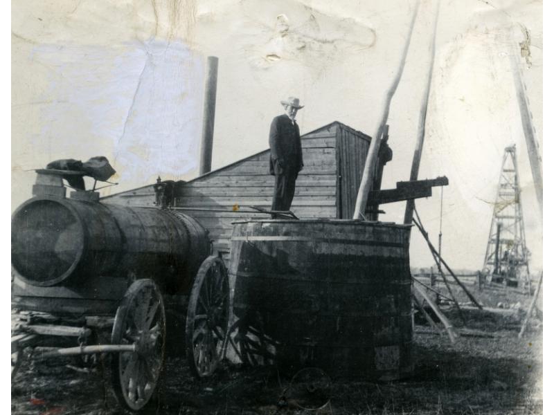 A man stands on top of a cylindrical wooden storage tank with metal rings around it. There is a boiler behind him and he is just to the side of a three-poled derrick. There is an oil rig in the background.