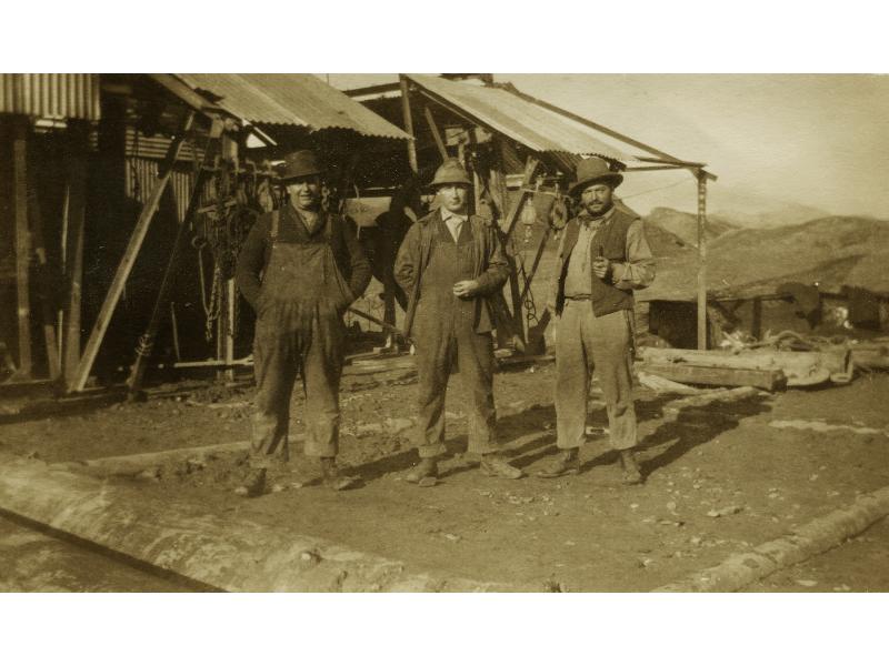 Three International Drillers wearing work clothes and hats, standing in front of an oil derrick with a roof made from metal sheeting. There are dirt hills in the background.