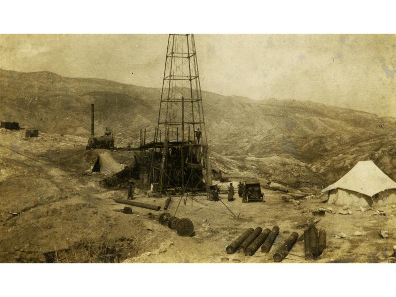 A metal oil rig. There are men standing around it and near a car. There are metal cylinders in the foreground and a tent on either side of the rig. There is a cylindrical steam boiler behind the rig. There are hills in the background.