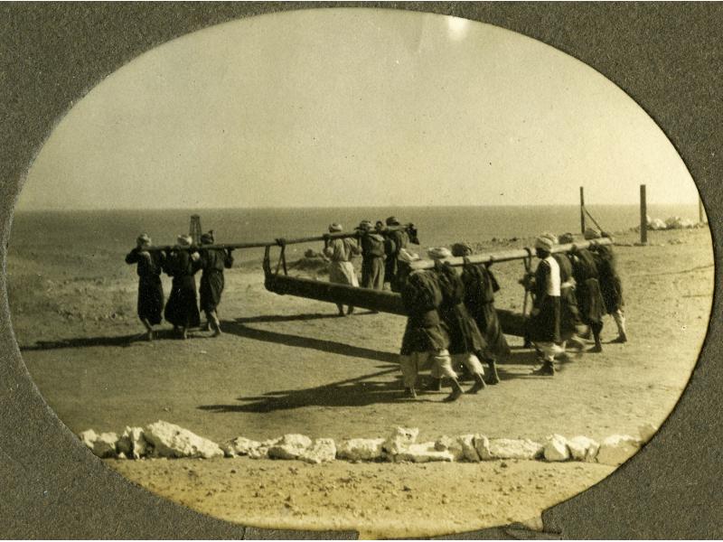 Une photo d’un groupe d’hommes transportant un tuyau. La conduite est fixée à deux poutres et les hommes se tiennent debout à l'avant et à l'arrière de la conduite et soutiennent la poutre sur leur dos. Le sol est plat et il y a une rangée de rochers blancs à l’arrière-plan.